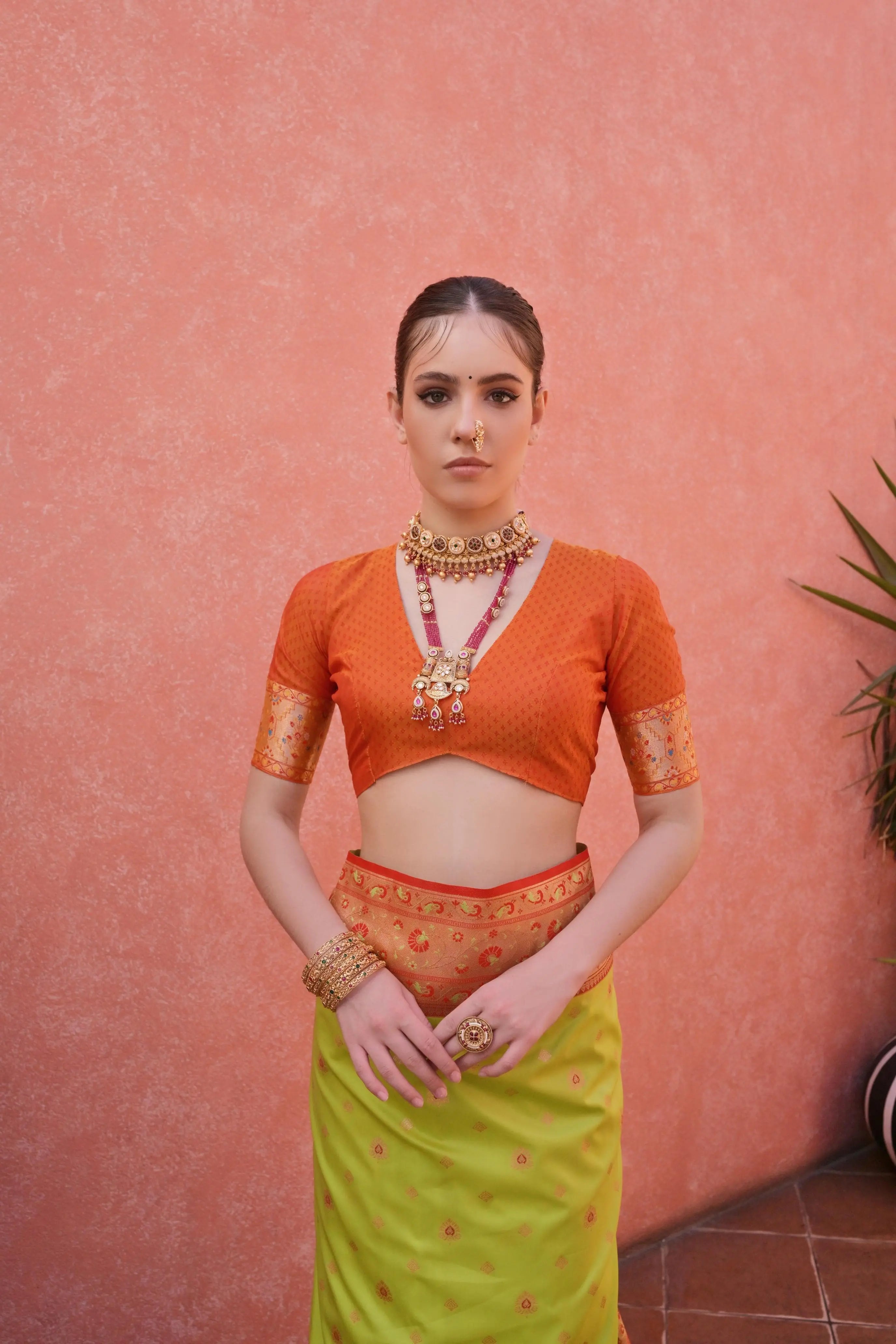 A woman models a vibrant Banarasi Silk Saree. The saree is lime green with fine Zari weaving, featuring a wide, ornate orange and red Zari woven border and pallu. She pairs it with a short-sleeve orange blouse and heavy traditional gold jewelry.